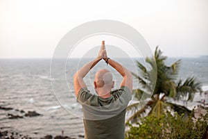 Rear view of vacationer folding palms together and lifting arms up, practicing yoga against seascape in front of sea.