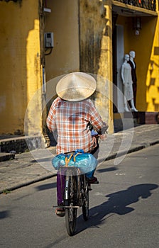 Rear view of a Vietnamese woman with a bamboo rice hat cycling in Hoi An old town