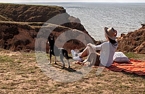 Rear View Of Woman With Dog Sitting On Hill Against Sea At Sunset.