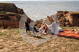 Rear View Of Woman With Dog Sitting On Hill Against Sea At Sunset.