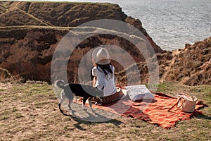 Rear View Of Woman With Dog Sitting On Hill Against Sea At Sunset.