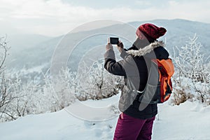 Rear view of a woman dressed warm taking pictures of snowy hills