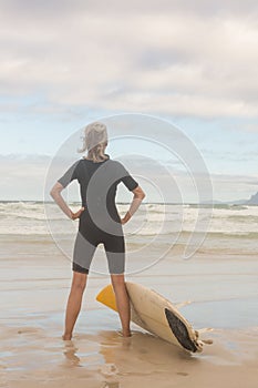 Rear view of woman with hand on hip standing by surfboard on shore