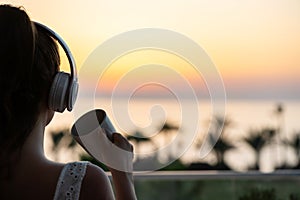 Rear view of woman with headphones and cup of tea looking on palm trees sea beach at sunset. Female relaxing listenning music.