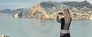 Rear view woman holding smartphone take photography of mountains hillside Calpe townscape and Mediterranean Sea. Capture moment,