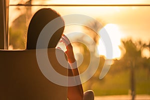 Rear view of woman sitting on chair near window and looking on palm trees sea beach at sunrise. Female relaxing.