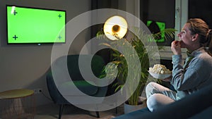 Rear view of a woman sitting on a sofa in the living room in the evening and watching a green TV screen mockup, is
