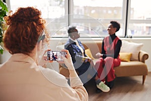 Rear view of woman taking photo on smartphone of two black female colleagues sitting on sofa by window