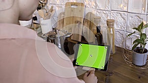 Rear view of a woman tapping on a tablet with green screen in the kitchen at home for recipes. Stock footage. Female