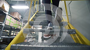 Rear view of a woman in uniform climbing up stairs inside warehouse. Creative. Storage center with goods on shelves.