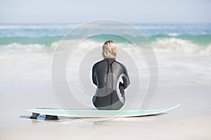 Rear view of woman in wetsuit sitting with surfboard on the beach