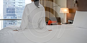 Rear view of woman in white bathrobe sitting on bed after waking up, looking at city view in the window. Copy space