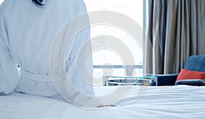 Rear view of woman in white bathrobe sitting on bed after waking up, looking at city view in the window. Copy space