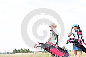 Rear view of women with golf club bags at course against clear sky