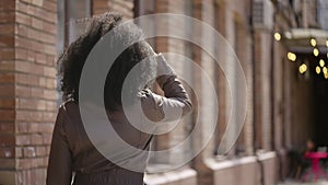 Rear view of young African American woman walking street, turning around and smiling. Brunette in brown leather jacket