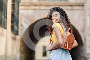Rear view of young Arab woman with backpack outdoors. Traveler girl in casual clothes in the street. Happy female wearing yellow t