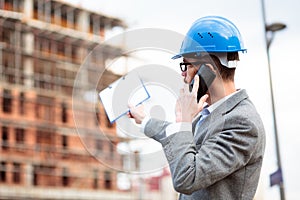 Rear view of a young architect or engineer inspecting construction site and talking on the phone