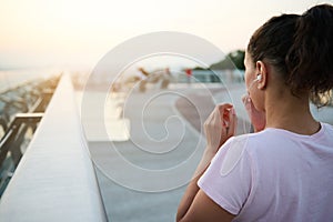 Rear view of a young athlete woman putting on earphones and getting ready for morning jog, looking at beautiful sunrise at the end