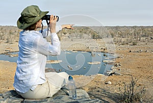 Rear view of a young blond woman on safari sitting on the rock looking through binoculars - travel concept
