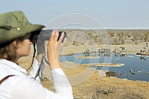 Rear view of a young blond woman on safari sitting on the rock looking through binoculars - travel concept