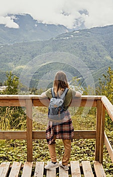 Rear view of young blonde woman with backpack and plaid shirt looking at view of Caucasian mountains and cloudy sky standing on