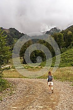 Rear view of young blonde woman with backpack walking uphill along trail on background of mountain covered with forest, active