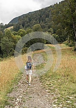 Rear view of young blonde woman with backpack walking uphill along trail on background of mountain covered with forest, active