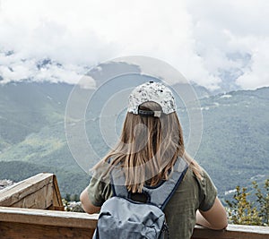 Rear view of young blonde woman in cap with backpack looking at view of Caucasian mountains and cloudy sky standing on wooden