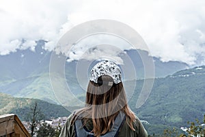 Rear view of young blonde woman in cap with backpack looking at view of Caucasian mountains and cloudy sky standing on wooden