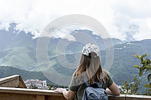 Rear view of young blonde woman in cap with backpack looking at view of Caucasian mountains and cloudy sky standing on wooden
