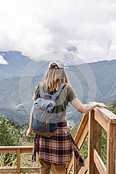 Rear view of young blonde woman in cap with backpack and plaid shirt looking at view of Caucasian mountains and cloudy sky