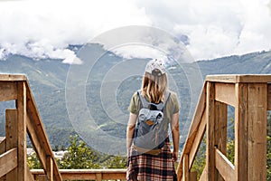 Rear view of young blonde woman in cap with backpack and plaid shirt looking at view of Caucasian mountains and cloudy sky
