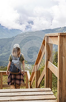 Rear view of young blonde woman in cap with backpack and plaid shirt looking at view of Caucasian mountains and cloudy sky