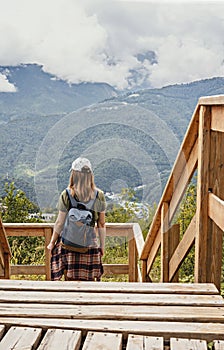 Rear view of young blonde woman in cap with backpack and plaid shirt looking at view of Caucasian mountains and cloudy sky