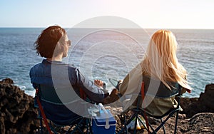 Rear View Of Young Couple Relaxing In Camping Chairs On Beach Rocks