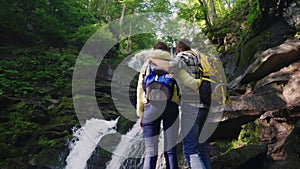 Rear view: A young couple of tourists with backpacks admiring the waterfall in the mountains. Stand on slippery stones