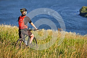 Rear view of the young cyclist stands with mountain bike on the green meadow above big river.