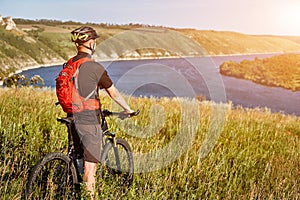 Rear view of the young cyclist stands with mountain bike on the green meadow above big river.