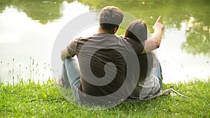 Rear view of young family sitting near the lake in the city park and kissing. Smiling happy man and woman spending time