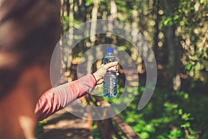 Rear view of young female holding bottle of fresh water in woods or park. Sport and recreation concept