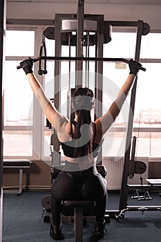 Rear view of young fitness lady in black sportswear sitting on gym machine and doing arm exercises with weights. A woman