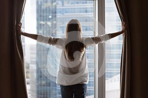 Rear view of young homeowner woman opening drapes at window