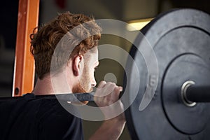 Rear View Of Young Man In Gym Lifting Weights On Barbell