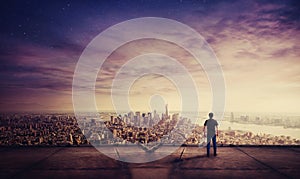 Rear view of young man standing on the rooftop of a skyscraper watching sunset over the big city horizon. Businessman on the roof