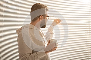 Rear view of young stylish man with beard and coffee peeking through hole in window blinds and looking out into street