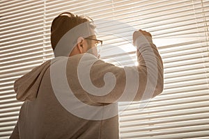 Rear view of young stylish man peeking through hole in window blinds and looking out into street. Concept of enjoying