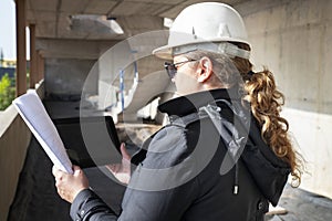 Rear view of young woman architect wearing a safety helmet, holding paper floor plans and checking a digital device