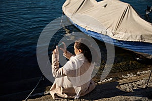 Rear view of a relaxed young woman in beige coat, sitting nears small boats by the Como lake and photographing the photo