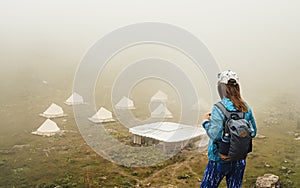 Rear view of young woman in cap and blue sportswear with backpack hiker standing in green mountain valley and looking at tent