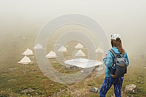 Rear view of young woman in cap and blue sportswear with backpack hiker standing in green mountain valley and looking at tent